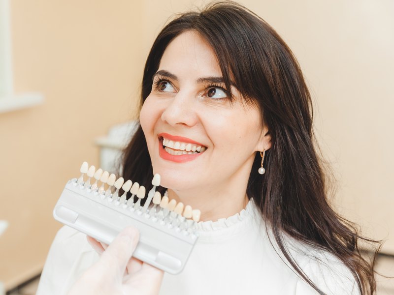 Young woman smiling and holding a whitening shade guide in beige and white tones, neutral background