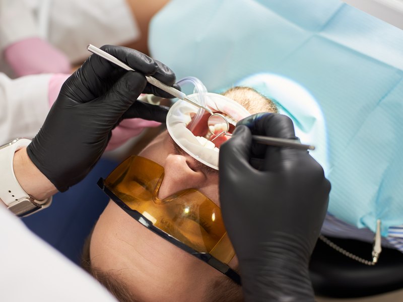 Dentist in black gloves working on a patient's tooth with a mirror and probe, patient wearing orange protective glasses