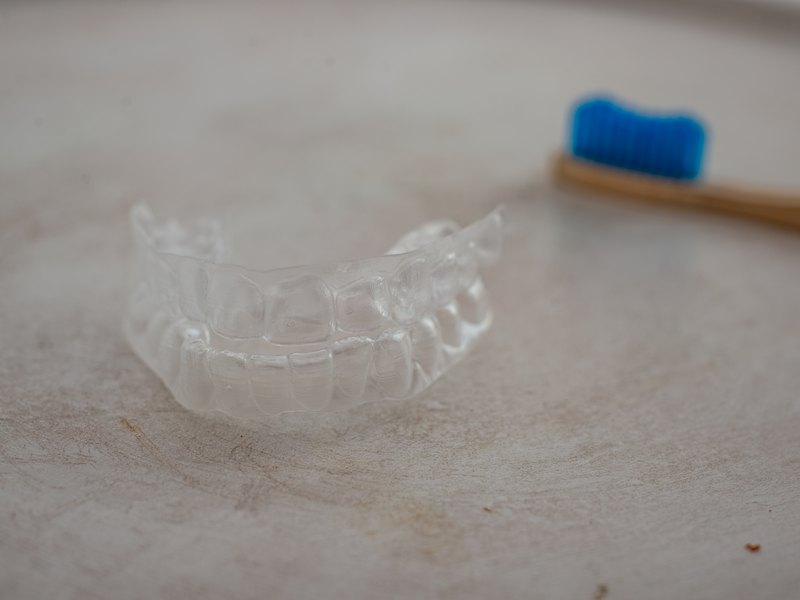 Clear orthodontic appliance on a plain surface, with a wooden-handled toothbrush behind