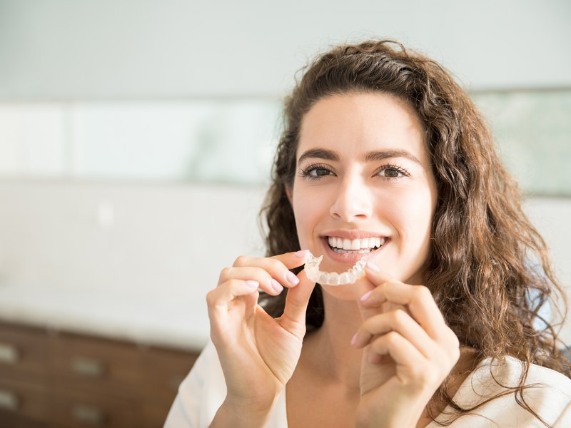 Woman holding a clear orthodontic aligner in her hands, smiling, plain white interior