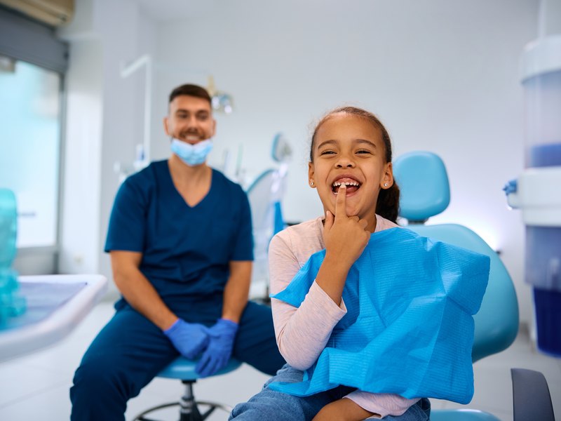 Dentist in a navy uniform smiling beside a young patient in a blue dental chair