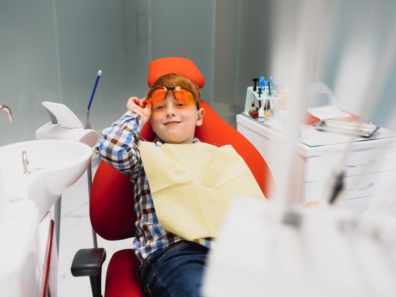 Child in a red dental chair with yellow protection, goggles and a protective cap