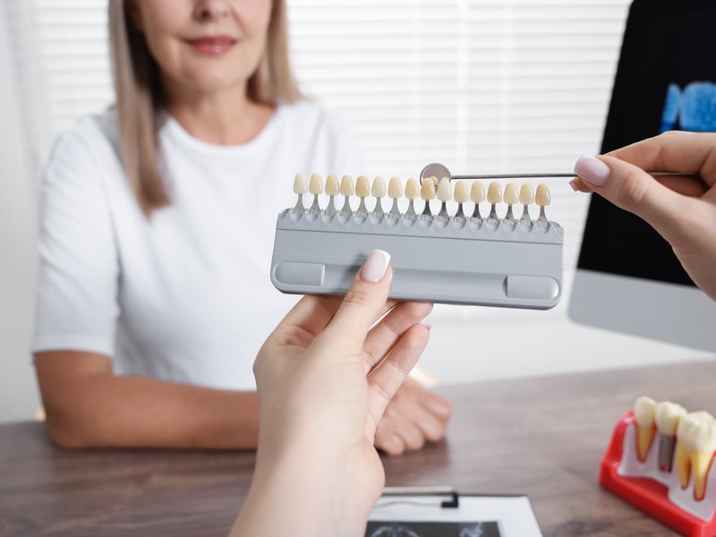 Two hands examining and comparing a dental shade guide, wooden table with dental material behind