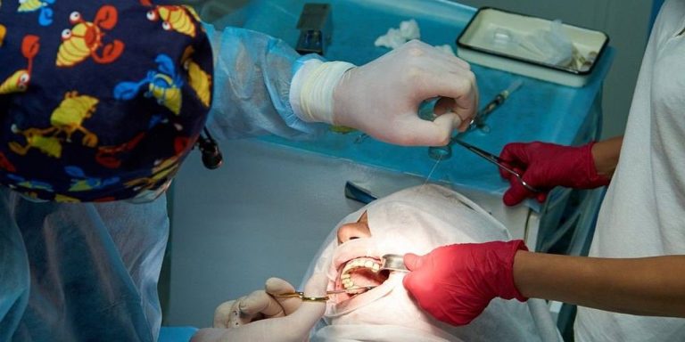 Dental surgeon placing a stitch in a patient's gum under a surgical drape, with an assistant in red gloves holding the lip back