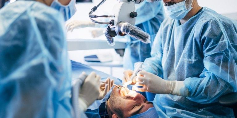 Dental surgeon looking through a microscope while operating on the mouth of a reclining patient, with an assistant beside him