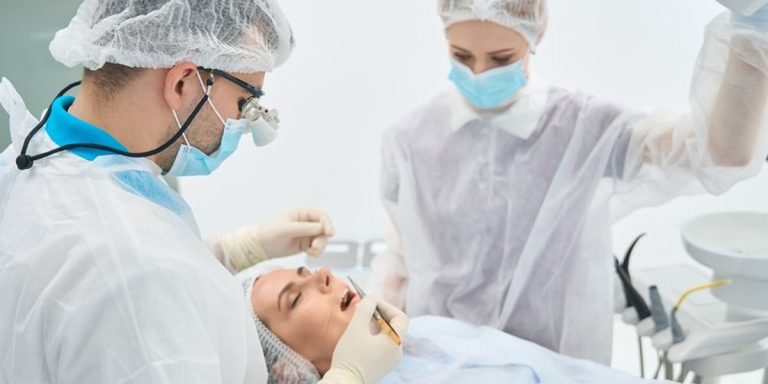 Cirujano con lupas y su ayudante, con gorro y mascarilla, operan la boca de una paciente tumbada en el sillón dental