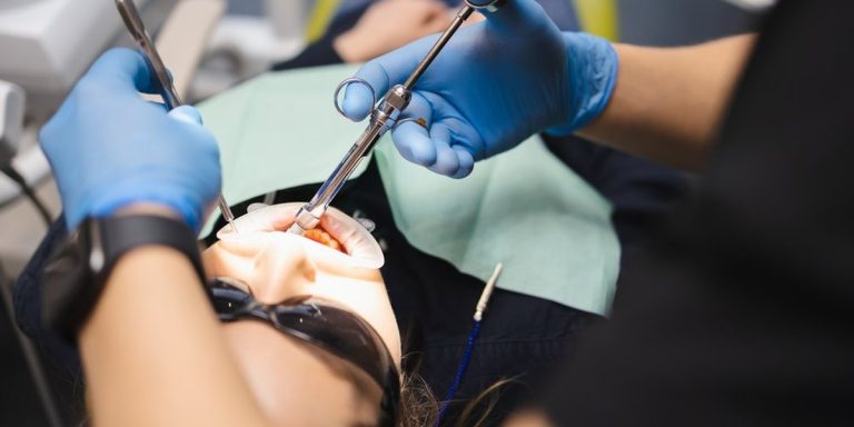 Dos manos con guantes azules trabajan con instrumental en la boca de una paciente tumbada en el sillón dental