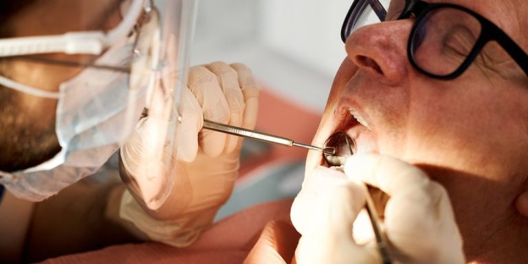 Dentista amb mascareta revisa amb mirall i sonda les genives d'un pacient amb ulleres que obre la boca
