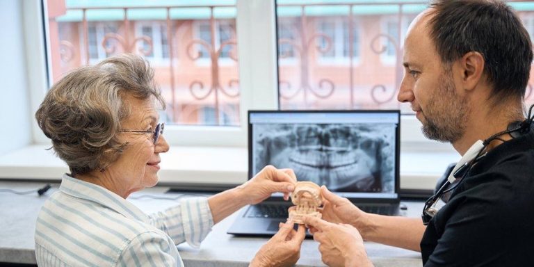 Pacient gran i dentista sostenen junts un model dental davant d'una radiografia panoràmica a l'ordinador portàtil