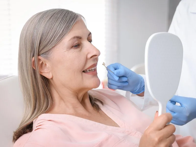 Senior woman checking her prosthetic teeth in a hand mirror while the dentist's gloved hand adjusts them in Barcelona